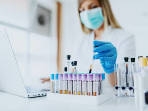 Close up of lab assistant in uniform, with mask and rubber gloves holding test tube with blood sample while typing on laptop